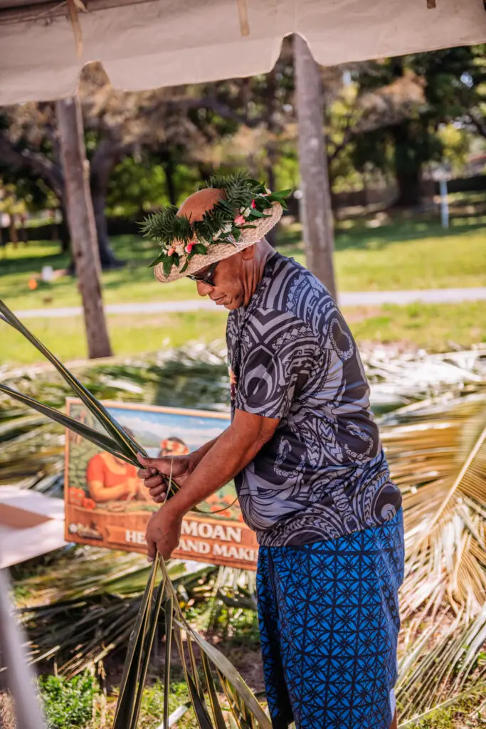 Island host demonstrating coconut palm leaf weaving at an outdoor event activity station