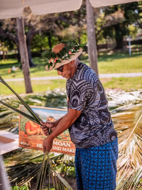 Island host demonstrating coconut palm leaf weaving at an outdoor event activity station